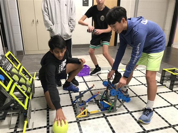 Students and their robot are on a VEX IQ competition field during practice testing their robot's scoring ability with a ball.